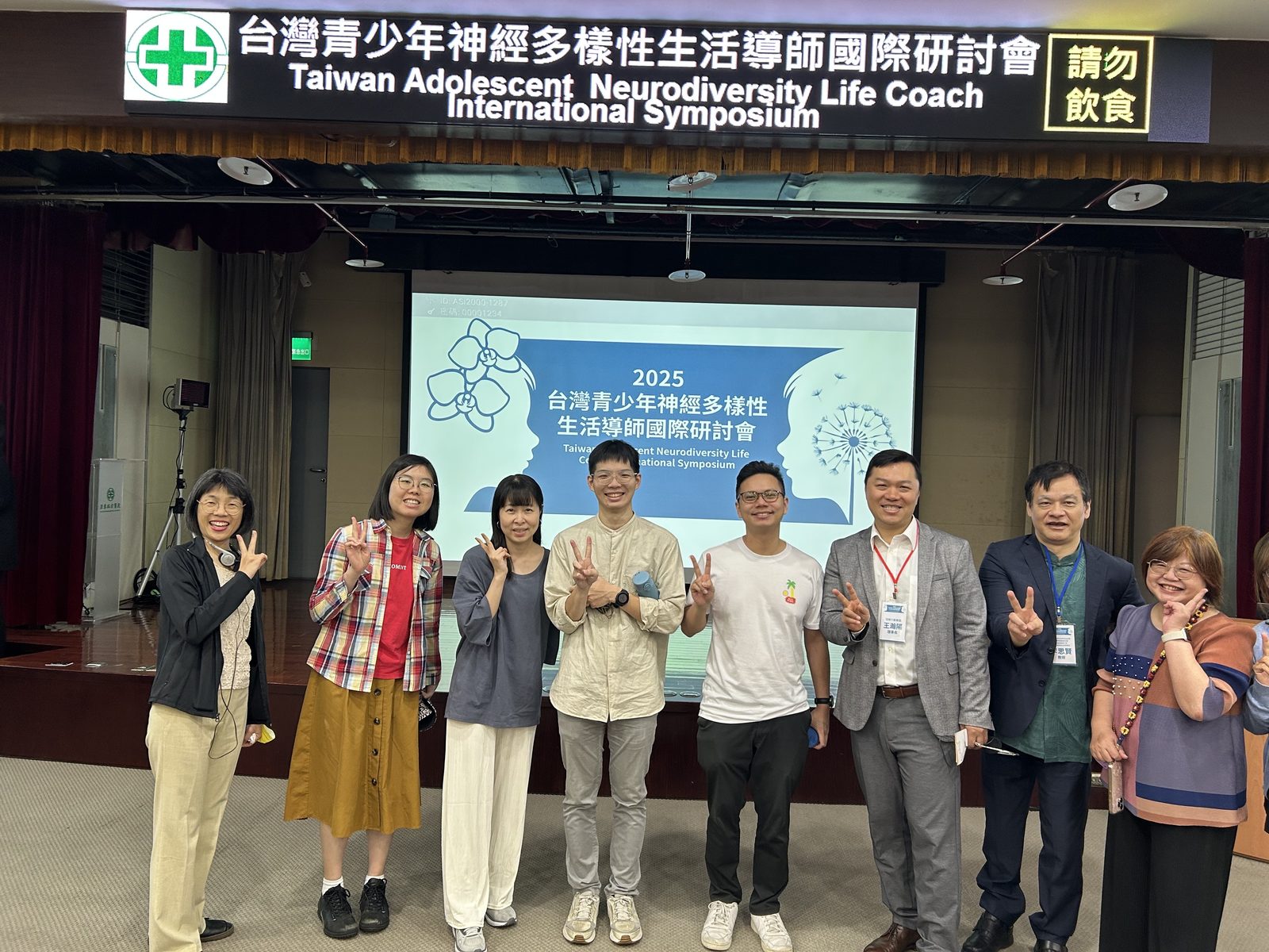 2025 年台灣青少年神經多樣性生活導師國際研討會現場，與會講者與工作團隊於主視覺投影幕前合影，投影片以蘭花與蒲公英為設計意象